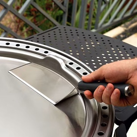 Hand using a stainless steel scraper tool with black handle to clean a silver BBQ grill surface outdoors