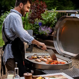 Man in apron cooking meat, fish and vegetables on a large round outdoor gas barbecue grill in a garden setting