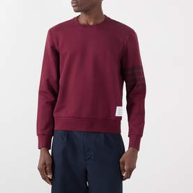 Model wearing Thom Browne burgundy cotton sweatshirt with tonal arm stripe detail and logo tab, styled with navy trousers.