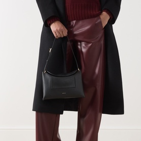 Model carrying Wandler Penelope Slouch Shoulder Bag in Black Crust, gold-toned hardware, styled with black coat and burgundy leather trousers.