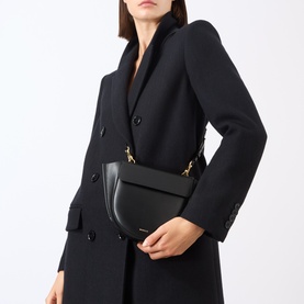 Model carrying Wandler Hortensia Mini Shoulder Bag in black calf leather with gold-toned hardware, worn crossbody over black coat.