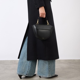Model carrying Wandler Hortensia Mini Shoulder Bag in black calf leather with gold-toned hardware, styled with black coat and wide-leg jeans.