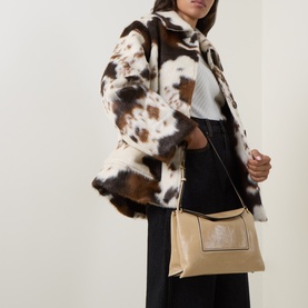 Model carrying Wandler Penelope Slouch Shoulder Bag in Sand Glaze pebbled leather, gold-toned hardware, adjustable strap, styled with cow-print coat