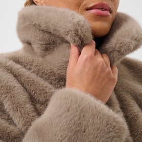Model wearing Kaffe Rosa Faux Fur Jacket in Taupe Gray, hands raised to soft stand collar, plush cocoon silhouette visible.