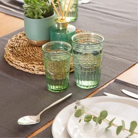 Two green embossed glass tumblers on a grey table runner, styled with woven placemat, cutlery, and eucalyptus sprig.