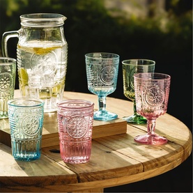 Set of multicoloured embossed glassware including goblets and tumblers in pink, blue and green, styled on a wooden table outdoors