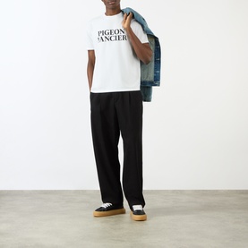 Model wearing Pellador white cotton T-shirt with "Pigeon Fancier" slogan, styled with black wide-leg trousers and platform trainers, denim jacket