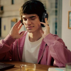 Model wearing black over-ear wireless headphones, adjusting fit, seated at desk wearing a pink hoodie