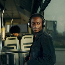 Model wearing black over-ear wireless headphones seated on a bus, styled with a dark jacket