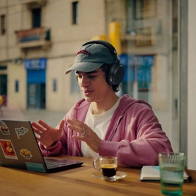 Model wearing black over-ear headphones at a caf&eacute; table with a laptop, dressed in a pink hoodie and grey cap