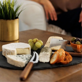 Swissmar walnut Knife Set displayed on slate board with cheese, grapes, olives and bread, gold plant pot in background
