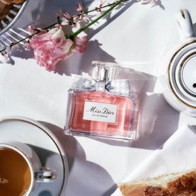 Dior Miss Dior Eau de Parfum bottle on white linen beside coffee cup, bread and pink flowers in bright natural light