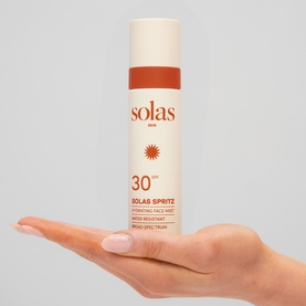 Model holding Solas Skin Solas Spritz SPF 30 Face Mist in white spray bottle with brown cap close to face