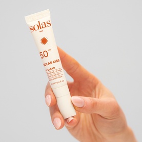 Solas Skin Kiss X Clear SPF 50 lip sunscreen tube held in hand, white packaging with sun graphic detail