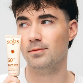 Model with Solas Skin Solas Sun SPF 50 sun cream tube, held against cheek, white packaging with gold sun motif