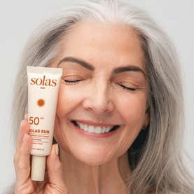 Model with Solas Skin Solas Sun SPF 50 tube held against cheek, cream/beige packaging with sun motif