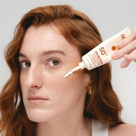 Model with Solas Skin SPF 50 sunscreen tube, applying product beneath eye area, white squeeze bottle with sun motif