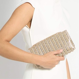 Model carrying Dune gold crystal-embellished clutch bag with stripe detailing, styled with a white sleeveless dress.