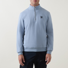 Model wearing Belstaff Mist Blue cotton half-zip sweatshirt with funnel neck, silver zip, and embroidered chest logo, styled with navy trousers.