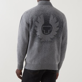 Model wearing grey knit zip-neck jumper with large intarsia Belstaff logo graphic on the back, paired with dark trousers.