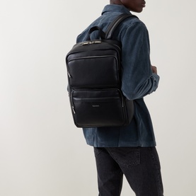 Model carrying Calvin Klein Raise Logo Dual Pocket Backpack in black with silver-tone hardware, styled with denim shirt and dark jeans