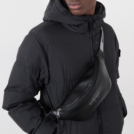 Model carrying Calvin Klein Bold Monogram Logo Bum Bag in black, worn crossbody, with zip fastening and logo detail, styled with black puffer jacket.