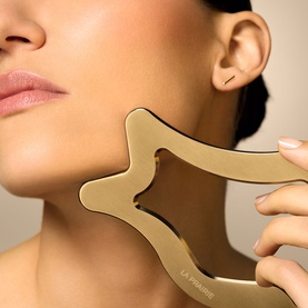 Model with La Prairie gold facial sculpting tool held against jawline and neck for firming and toning
