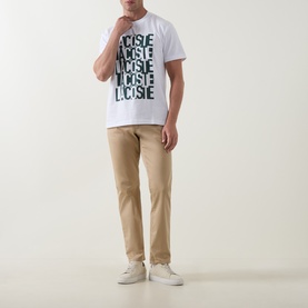 Model wearing Lacoste white crew neck T-shirt with repeated green logo print, styled with beige chinos and white trainers.
