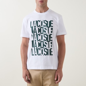 Model wearing Lacoste white cotton crew neck T-shirt with repeated bold green Lacoste logo print, styled with beige trousers.
