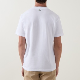 Model wearing Lacoste Crew Neck T in white, rear view, with green crocodile logo at nape, styled with beige chinos.