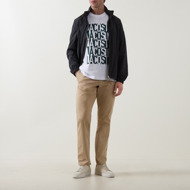 Model wearing Lacoste white crew neck T-shirt with repeated logo print, styled with black jacket and beige chinos.