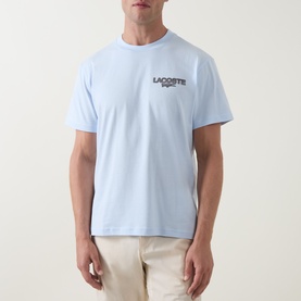 Model wearing Lacoste Crew Neck T in light blue with small chest logo graphic, styled with beige trousers.