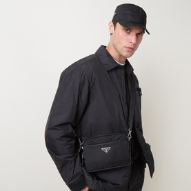 Model carrying Prada Bandoleria Triangle Logo Crossbody Bag in black with silver triangle logo, worn crossbody over black jacket