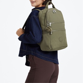 Model carrying Kipling backpack in Almost Sage, featuring top handle, zip pockets, and signature monkey keychain, styled with denim jacket.