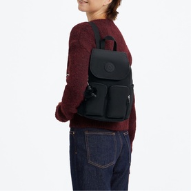 Model carrying a small black backpack with front zip pockets, flap closure, and black hardware, styled with a red knit jumper and jeans