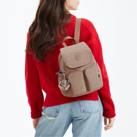 Model carrying a small taupe nylon backpack with flap closure, monkey keychain, and front pockets, styled with a red knit top and jeans