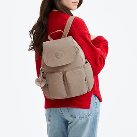 Model carrying a taupe crinkle nylon backpack with front pockets, flap closure, and monkey keychain charm, styled with a red sweater and jeans.