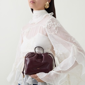 Model carrying Chloe Icons Leather Mini Crossbody Bag in Burnt Red with drawstring closure and top handle, styled with white lace blouse and gold