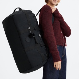 Model carrying large black duffle bag with silver hardware and shoulder strap, styled with red knit jumper and jeans