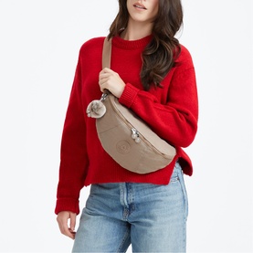 Model carrying a taupe crescent crossbody bag with pom-pom charm and gold-tone zip, worn over a red sweatshirt with jeans.