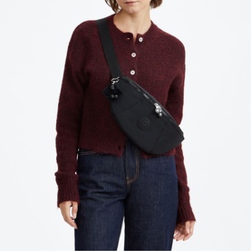 Model carrying a black Kipling belt bag with silver-tone hardware, worn crossbody, styled with a dark red knit and jeans