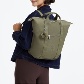 Model carrying large green Kipling tote backpack with silver hardware and signature monkey keychain, styled with navy jacket and brown trousers