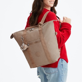 Model carrying a large sandy beige Kipling tote bag with zip top, adjustable strap and signature monkey keyring, styled with red sweater and jeans