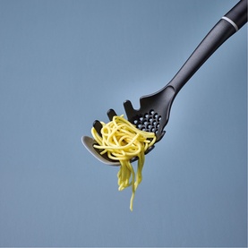 Prestige grey spaghetti spoon with perforated head and tong-style prongs, shown holding cooked pasta against a blue background