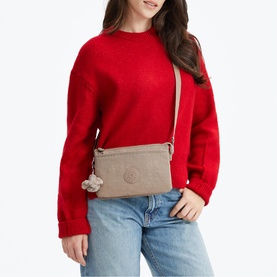 Model carrying a small taupe crossbody bag with long adjustable strap and Kipling monkey charm, styled with red sweatshirt and jeans