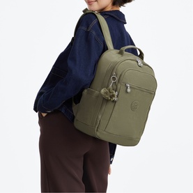 Model carrying an olive green Kipling backpack with top handle, silver-tone zips, and signature monkey keyring charm