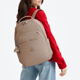 Model carrying a large beige Kipling backpack with multiple zip compartments, worn over one shoulder, styled with a red sweater and jeans.