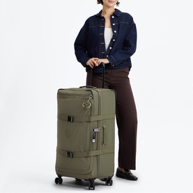 Model carrying large olive green soft-shell suitcase with spinner wheels and top handle, styled with denim jacket and brown trousers