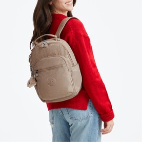 Model carrying a beige Kipling backpack with silver hardware, multiple zip pockets and hanging monkey charm, styled with red jumper