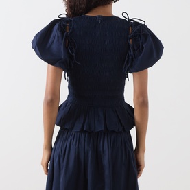 Model wearing Damson Madder navy cotton blouse, back view, with puff sleeves, ribbon tie details, smocked bodice and peplum hem.